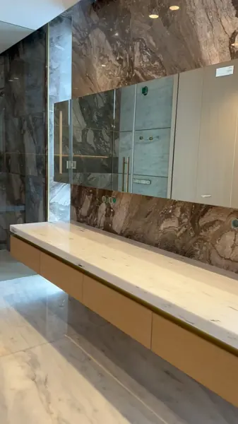 Warm beige natural stone is used for the vanity and its surrounding surfaces, alongside warm metallic details and carefully placed lighting.