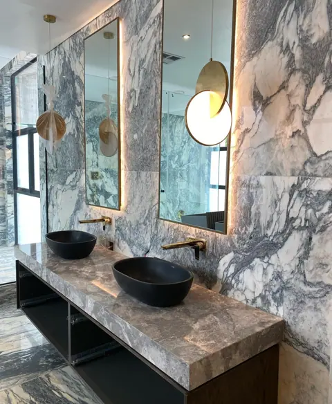 Stone with mirrored veining is used for the vanity and its surrounding surfaces, alongside timber cabinetry and a round mirror.