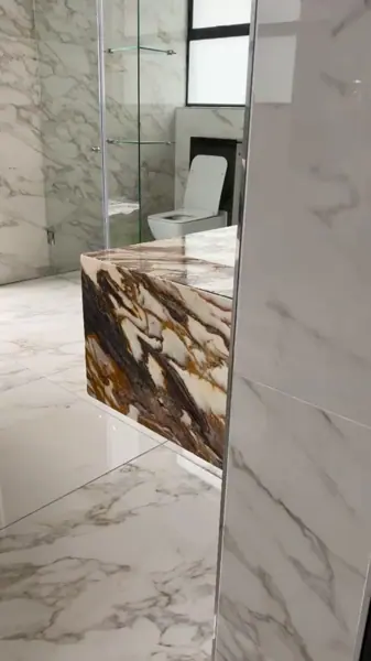 Rich brown veined stone is used for the vanity and its surrounding surfaces, alongside tall windows and a bright neutral interior.