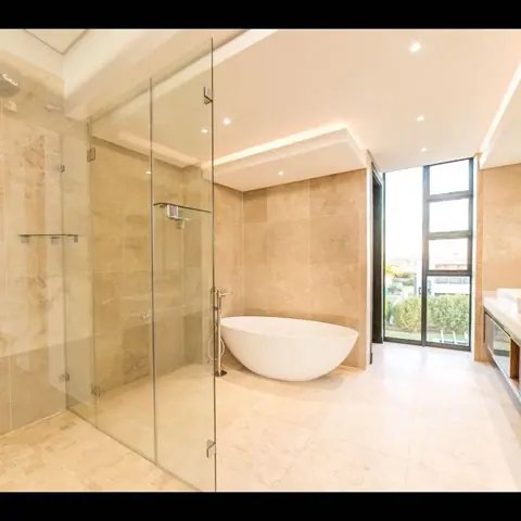 Warm beige natural stone is used for the bathroom walls and floor, alongside a freestanding white bath and soft lighting.