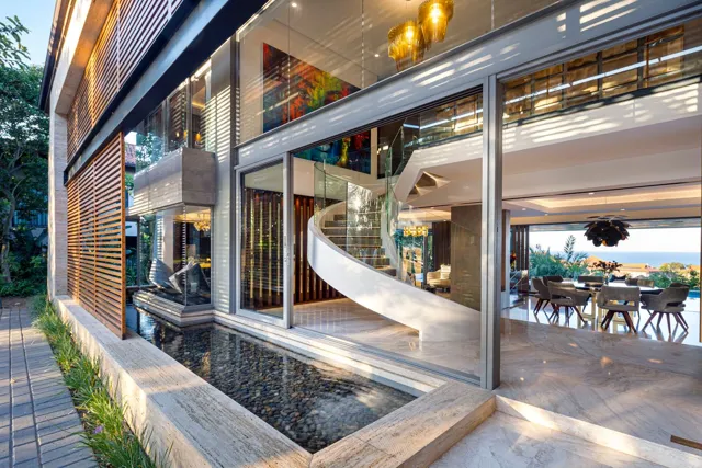 Broad glazed openings connect pale stone flooring, an outdoor water feature and a sculptural staircase in this open living space.
