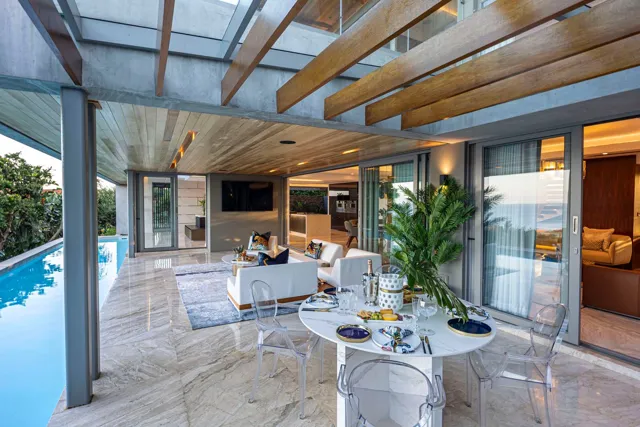 Warm stone flooring runs beneath a timber and glass canopy, framing a relaxed dining and seating area beside the swimming pool.