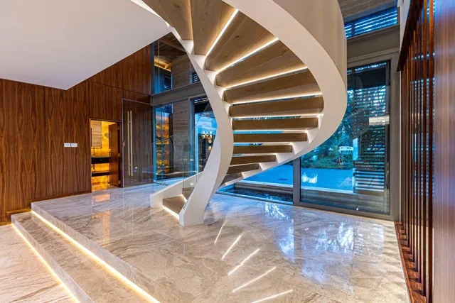 An illuminated curved staircase rises above polished stone flooring, where natural veining and reflected light emphasise the entrance hall.