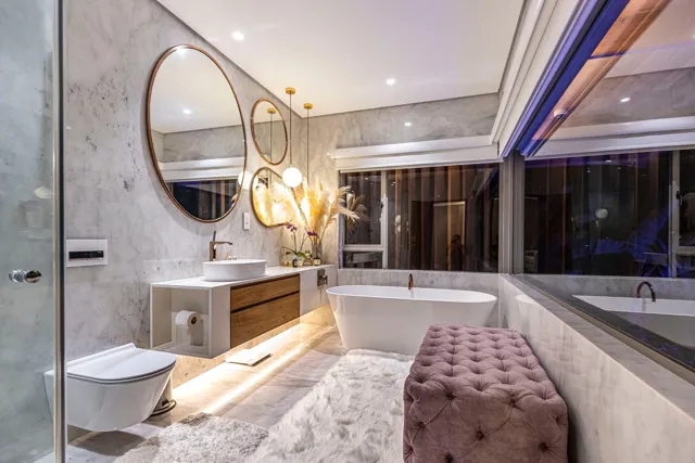 Pale stone walls and flooring surround a double vanity and inset bath, with warm lighting adding depth beneath the cabinetry.